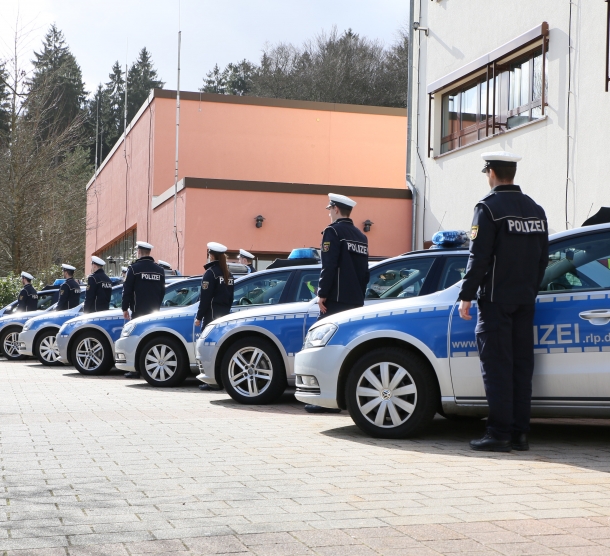 Sieben blau-silberne Polizeiwagen stehen angewinkelt nebeneinander. Neben jedem Wagen steht ein Polizist.