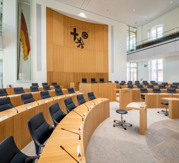 Bild des Plenarsaals des landtags Theinland-Pflaz. Alle Stühle sind leer. Im Hintergrund das Wappen des Landss Rheinland-Pfalz.