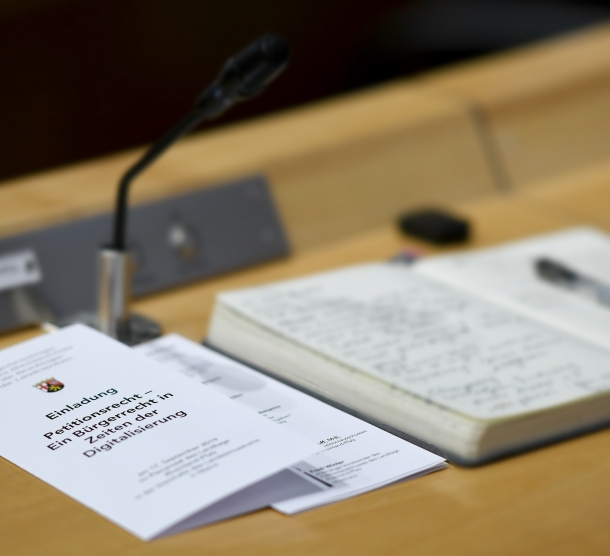An einem Tisch im Plenarsaal des Landtags liegt ein Notizbuch mit handschriftlichen Notizen. Daneben liegt eine Einladung zu einer Veranstaltung zum Petitionsrecht.