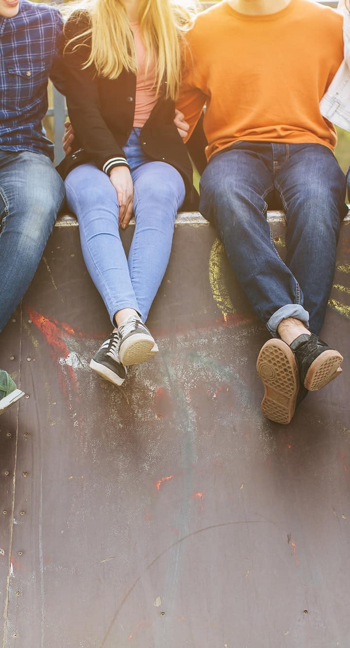 Auf einer grauen Mauer sitzen sechs Kinder und Jugendliche in Jeans, deren Beine locker und über Kreuz an der Mauer baumeln.